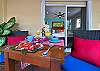 Private balcony at Abbey del Sol A2 Unit 10, a Puerto Morelos vacation rental, featuring a table with colorful place settings, red cushions, tropical plants, and a view back into the apartment.