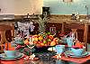 Dining area inside Abbey del Sol A2 Unit 10, a Puerto Morelos vacation rental, featuring colorful place settings, fresh fruit, wood cabinetry, and the kitchen in the background for an easy stay.