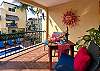 Private balcony at Abbey del Sol A2 Unit 10, a Puerto Morelos vacation rental, featuring a table with colorful settings, red cushions, tropical plants, wall decor, and a view over the pool and courtyard.