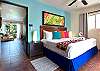 Bedroom inside Abbey del Sol A2 Unit 10, a Puerto Morelos vacation rental, featuring a king bed, colorful pillows, wood headboard, air conditioning, natural light, and an open view toward the living area.