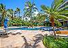 A wider view shows the shared free-form pool, stone deck, lounge chairs, umbrellas, and surrounding palms, highlighting the landscaped recreation area available to guests of The Watermark Waikiki on extended stays.