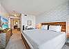 A reverse bedroom angle shows the king bed, long dresser, large flat-screen television, dedicated desk, bedside lighting, and en-suite access—an extended-stay setup that supports rest, storage, work, and privacy.