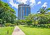 This garden-level exterior view places The Watermark Waikiki tower beyond a broad lawn, walking path, palms, and mature shade trees, highlighting the landscaped setting around the 35th-floor residence in Honolulu.