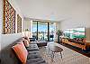The living room pairs a spacious sectional and large flat-screen television with direct lanai/balcony access, while floor-to-ceiling glass brings ocean, marina, and Honolulu skyline views into the central gathering space.