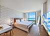 The primary bedroom combines a king bed with a dedicated desk, large flat-screen television, reading lamps, and a floor-to-ceiling window overlooking the Pacific Ocean—well suited to rest and remote-work routines.