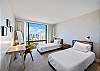 The guest bedroom provides two twin beds, individual reading lights, a shared nightstand, and a dedicated desk beside a large window—flexible sleeping and workspace arrangements for an extended Waikiki stay.