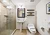 The guest bathroom includes a glass-enclosed walk-in shower, pedestal sink, open wall shelving, and fresh towel storage, giving occupants of the second bedroom a practical separate bath for longer stays in Waikiki.