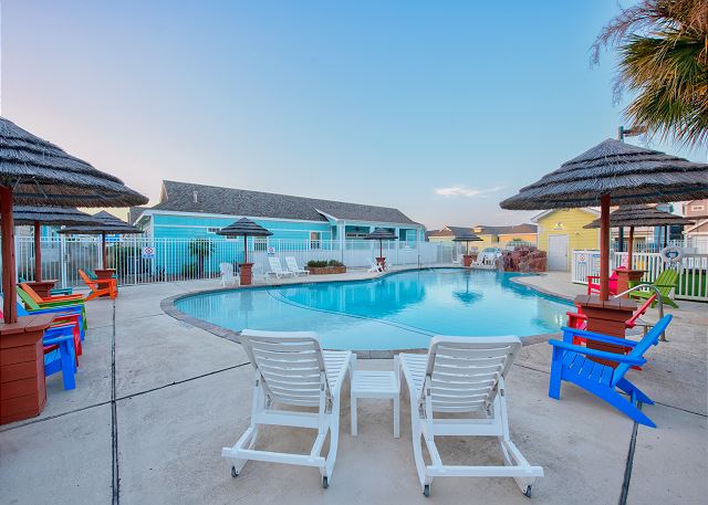 Inviting community pool with clear blue water, surrounded by comfy loungers and straw covered structures.