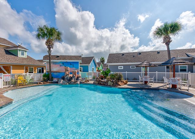 Inviting community pool with clear blue water, surrounded by comfy loungers and straw covered structures.