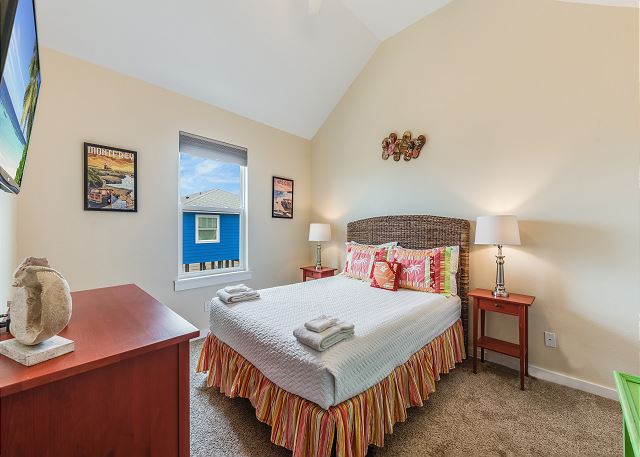 This cozy bedroom features a queen-sized bed with vibrant bedding, soft lighting from bedside lamps, and a window offering a view of charming coastal homes.