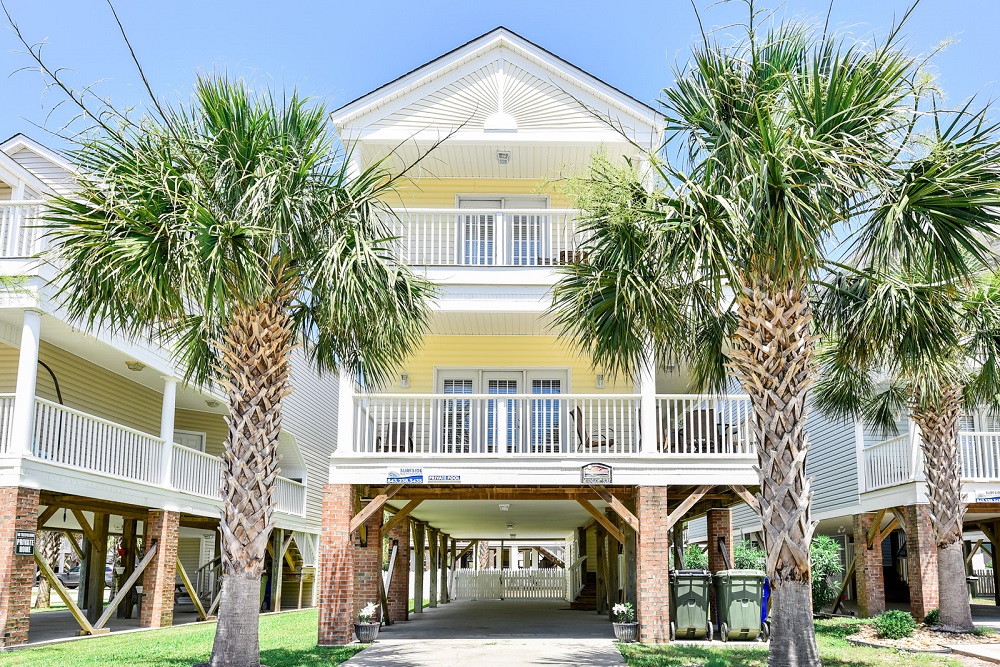 A photo of the front of a two story house in Surfside City, NC. The house is pale yellow with white trim and is framed by two palm trees.