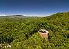 Wide aerial shot of the cabin surrounded by dense mountain forest under a clear blue sky.