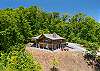 Full two-story cabin exterior with wraparound deck, hot tub, fire pit, and gravel parking. 