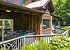 Covered deck area with a private hot tub, wood siding backdrop, and greenery on all sides.